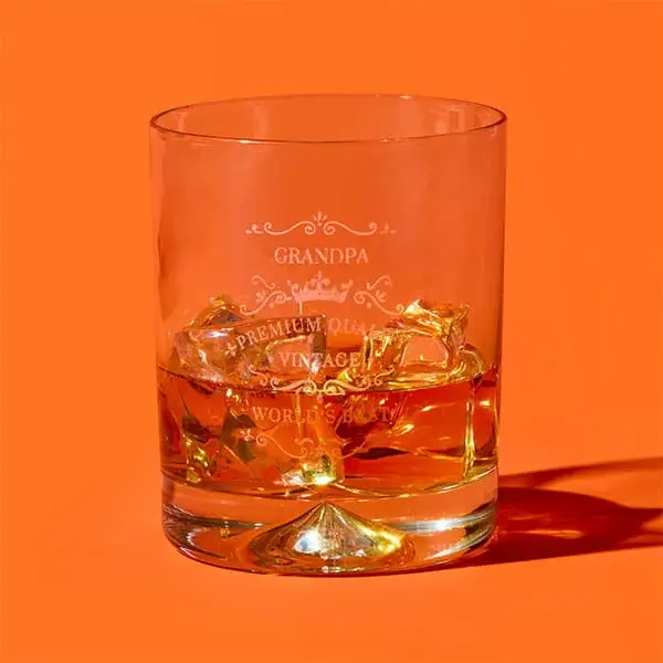 A personalised whisky tumbler engraved with a Grandpa design, filled with amber liquid and ice cubes, displayed against a bright orange background.